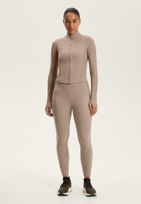 Frau steht frontal in einer beigefarbenen, enganliegenden Zip-Jacke, passenden Leggings und schwarzen Sportschuhen vor einem einfarbigen Hintergrund.