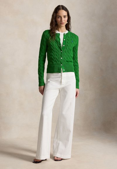 Žena v zelenom svetríku so zapínaním na gombíky cez biely top, v kombinácii s bielymi širokými nohavicami s vysokým pásom a čiernymi sandálmi.