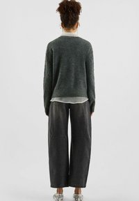 Persona con capelli ricci raccolti in uno chignon alto, che indossa un maglione grigio a trecce, una camicia bianca trasparente, jeans neri a gamba larga e scarpe con motivo, di spalle.