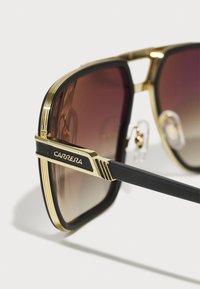 ochelari de soare Carrera cu ramă din plastic negru, accente metalice de aur, lentile maro cu gradient și pernuțe pentru nas ajustabile.
