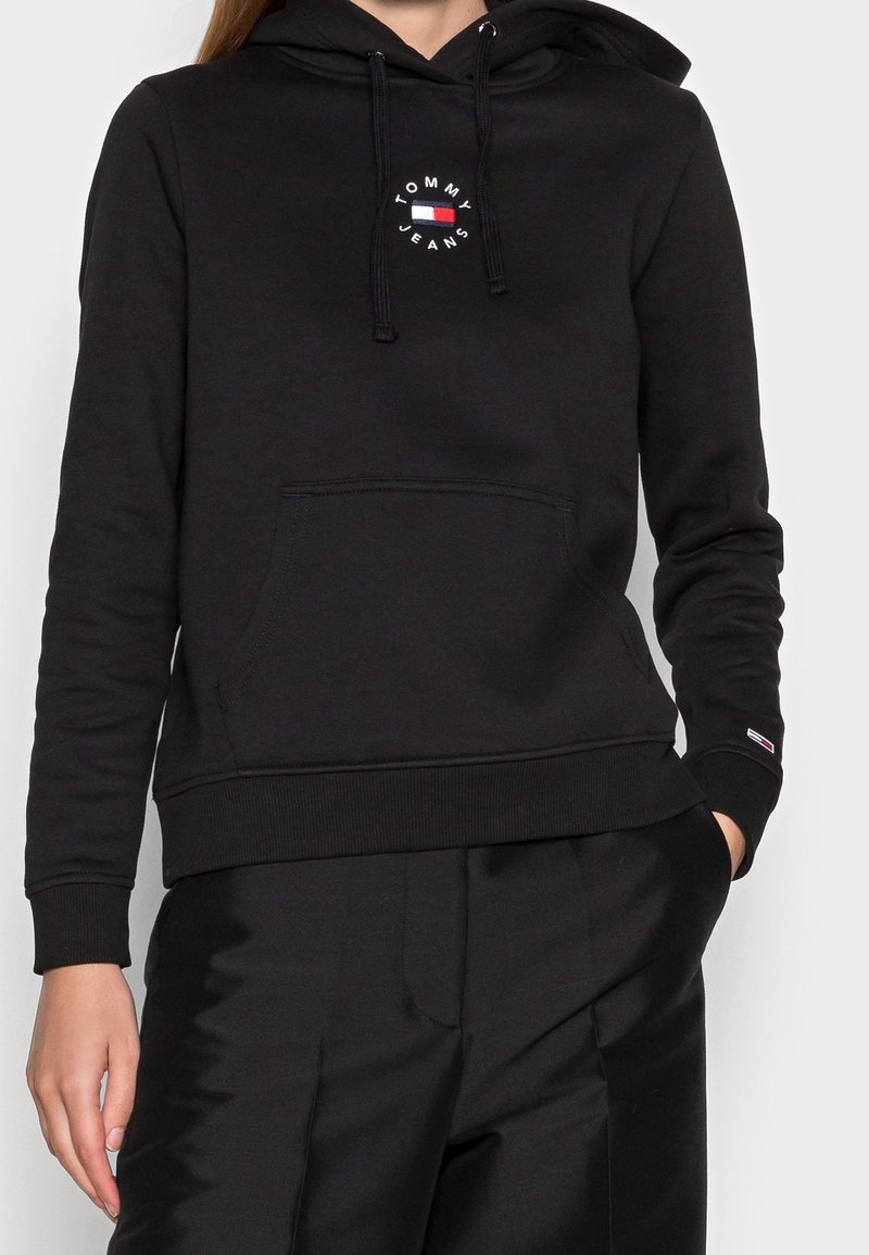 Černá bavlněná mikina s kapucí, přední kapsou a kruhovým logem s červenými a modrými akcenty na hrudi.