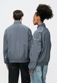 To personer i grå jakker med hvit piping, med høy krage og elastiske mansjetter, stående rygg mot rygg, iført mørke bukser.