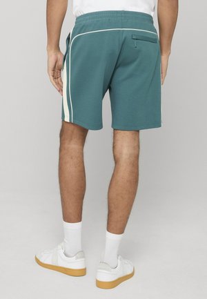 Man draagt turquoise sportkorte broek met witte zijstrepen, witte sokken en witte sneakers met gumzolen, staand met de rug naar de camera tegen een grijze achtergrond.