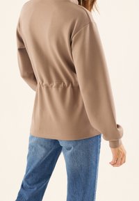 Jasnobrązowy sweter z ściągaczem w talii, długimi rękawami i miękką fakturą. Zestawiony z luźnymi niebieskimi dżinsami. Neutralne tło.