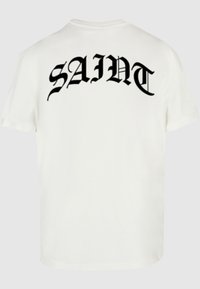 Hvid bomulds-t-shirt med fed, sort gotisk tekst "SAINT" trykt på ryggen. Standard rund hals og korte ærmer.