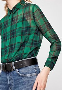Camisa de cuadros verdes con botones, hecha de material translúcido, que presenta un collar, mangas largas y detalles en negro en los puños. Se lleva con jeans de mezclilla.