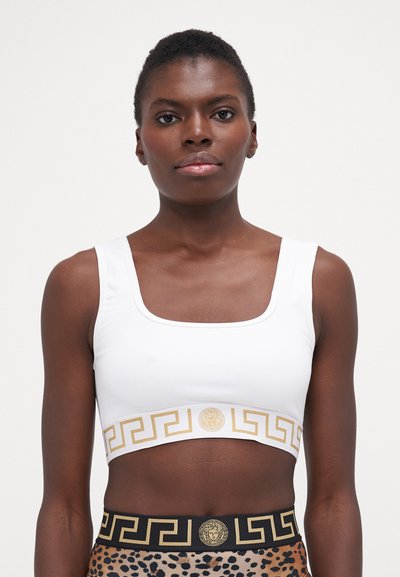 Bílý crop top s čtvercovým výstřihem a zlatým řeckým vzorem na lemu, v kombinaci s černým a zlatým vzorovaným páskem.