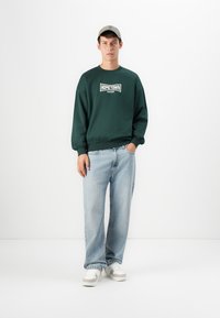 Young man wearing a green "HOMETOWN" sweatshirt, light blue jeans, beige cap, and white sneakers, standing with one hand in pocket.