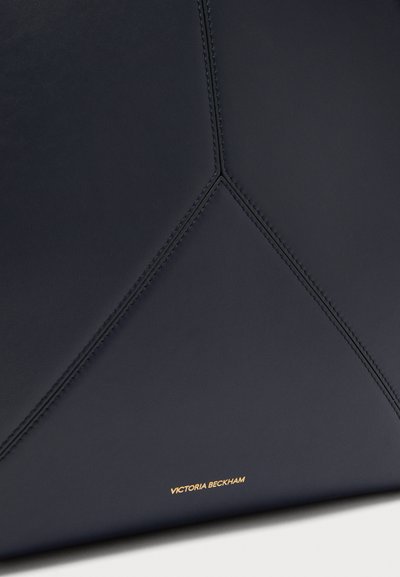 Εικόνα κοντινού πλάνου μαύρης δερμάτινης τσάντας με γωνιακό ραμμένο σχέδιο και μικρό χρυσό λογότυπο "Victoria Beckham" κοντά στην κάτω άκρη.
