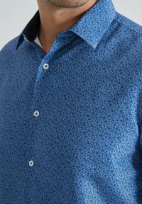 Chemise bleue à manches longues avec un petit motif floral, un col contrastant et une texture lisse. Elle présente des boutons blancs avec des centres foncés.