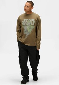 Maglietta a maniche lunghe color oliva con un design grafico, che presenta il testo "Star Wars" e una stampa di personaggi. Pantaloni cargo neri in tessuto testurizzato e sneakers.