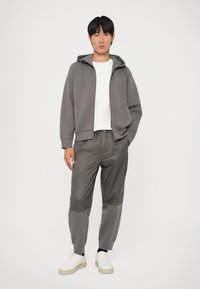 Felpa grigia con cappuccio e tasche frontali, abbinata a jogger grigi a gamba affusolata con pannelli a contrasto. Sneakers bianche completano il look.