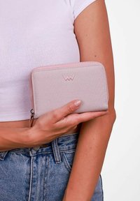 Světle růžová peněženka vyrobená z texturované tkaniny, s bočním uzávěrem na zip a vyraženým logem na přední straně. Držena v ruce osoby.