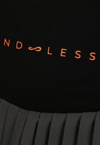 Tessuto nero con testo arancione "ND∞LESS" che presenta un simbolo dell'infinito al posto della lettera "O", sopra un indumento grigio plissettato.