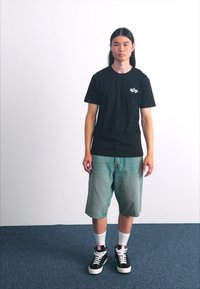 Schwarzes Baumwoll-T-Shirt mit einem kleinen weißen Grafik, kombiniert mit hellblauen Jeansshorts, weißen Socken und schwarzen Turnschuhen mit weißen Akzenten.