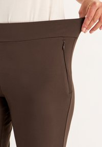 Braune Yogahosen aus deastischem Stoff, mit einem breiten Bund und einer unauffälligen Reißverschlusstasche an der Seite. Glatte Textur.