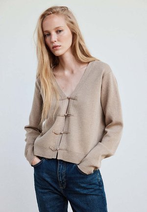 Junge Frau mit langen blonden Haaren, die eine beige Strickjacke mit Knebelverschlüssen und blaue Jeans trägt, steht mit den Händen in den Taschen.