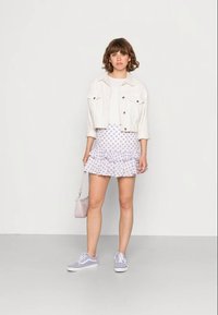 Jovem mulher em pé, vestindo uma jaqueta branca cropped, blusa branca, saia rufada floral roxa, ténis lilás e segurando uma pequena bolsa lilás.