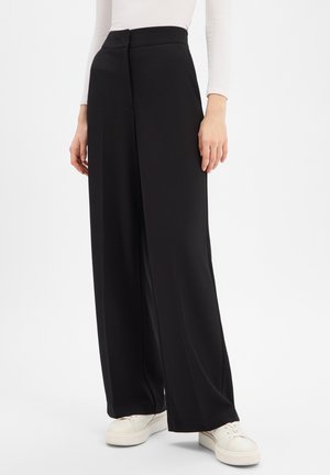 Schwarze Hose mit weitem Bein aus glattem Stoff, mit hoher Taille und einem vorderen Verschluss, kombiniert mit weißen Plateausneakers.