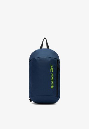 Granatowa torba na ramię Reebok z pionową kieszenią na zamek i limonkowym logo, czarna rączka na górze, stojąca pionowo na białym tle.