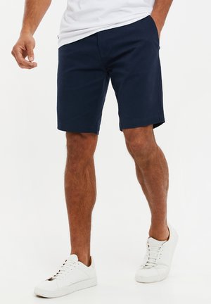 Marineblaue Shorts mit flacher Front aus Baumwolle, mit glatter Textur, Seitentaschen und abgestepptem Saum, kombiniert mit weißen Sneakers.