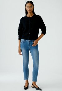 Femme se tenant debout dans un jean skinny bleu clair, un cardigan noir à boutons et des escarpins noirs à bout pointu devant un fond clair.