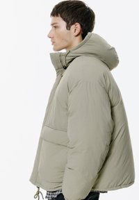 Svetlozelená puffrová bunda s kapucňou, s textúrovaným povrchom, elastickými šnúrkami a veľkým predným vreckom. Bočný pohľad.