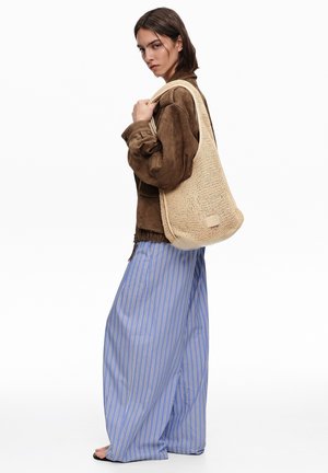 Mujer con chaqueta marrón de ante y pantalones anchos de rayas azules, llevando un gran bolso de hombro beige tejido.