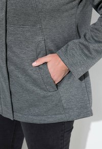 Handen i fickan på en grå texturerad jacka bärs med mörka byxor, mot en enfärgad ljus bakgrund.