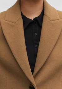 Bruine wolmix jas met geknipte revers en een enkele knoopsluiting, gedragen over een zwarte geribbelde shirt met drie knopen.