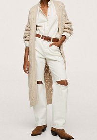 Beżowy dziergany kardigan, biała koszula, białe podniszczone dżinsy z przetarciami, brązowy pasek i brązowe botki na obcasie. Widoczny fakturowany wzór dzianiny.