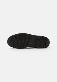 Semelle de chaussure en caoutchouc noir avec un motif texturé pour l'adhérence, arborant un accent vert et un logo sur le côté. Design durable et robuste.