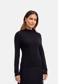 Frau mit glattem braunem Haar, die einen enganliegenden schwarzen Pullover mit langen Ärmeln und einen schwarzen Rock trägt, steht vor einem schlichten hellen Hintergrund.