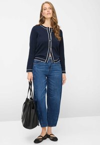 Marineblå cardigan med hvid kant, knaplukning, stylet med blå vidde jeans. Modellen holder en sort håndtaske og bærer sorte net-sko.