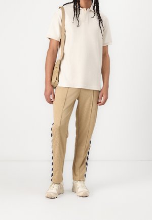 Personne portant un pantalon de survêtement beige avec des bandes latérales foncées, un polo crème, des baskets crème et portant un sac à bandoulière beige.