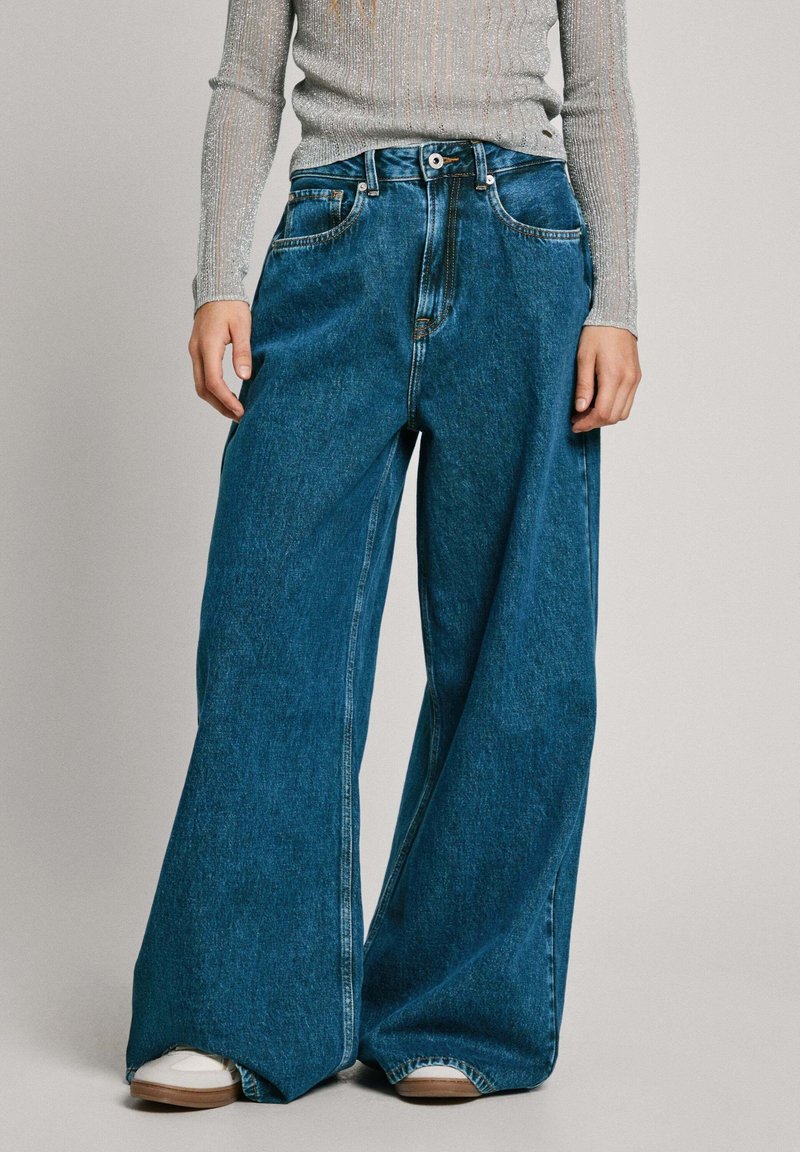 Personne portant un jean denim bleu à jambes larges avec une taille boutonnée, associé à des chaussures blanches et un haut gris à manches longues.