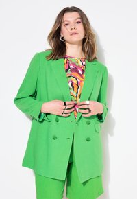 Dubbelknäppt grön blazer i lättviktsmaterial, matchad med en flerfärgad mönstrad topp och gröna shorts. Accessoarer inkluderar silverringar.