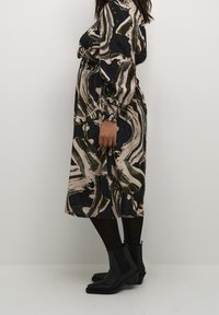 Robe midi à motif dans des teintes sombres avec des tourbillons abstraits de vert, beige et noir ; manches longues ; associée à des bottines noires.