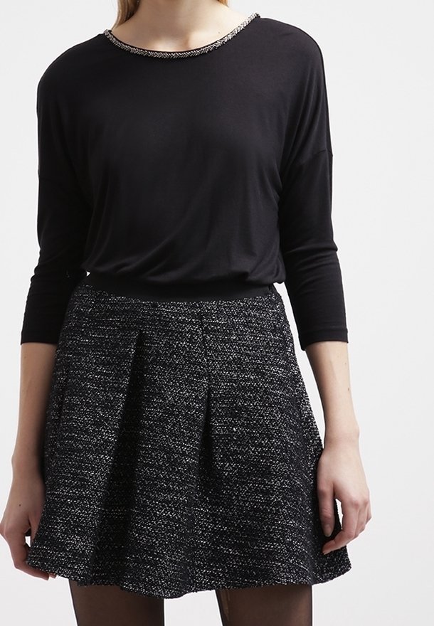 Femme portant un haut noir à manches longues avec un col orné de perles et une jupe plissée noire et blanche texturée.