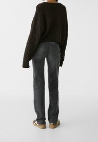 Suéter de punto marrón oscuro con diseño de trenzas, combinado con jeans de mezclilla grises y zapatillas marrones con acentos claros. Vista trasera.