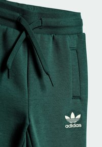 Zielone sportowe spodnie dresowe wykonane z teksturowanej tkaniny, z regulowanym sznurkiem w pasie, bocznymi kieszeniami oraz białym logo Adidas na przodzie.