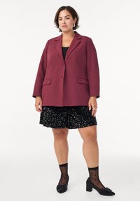 Kvinde iført en bordeauxfarvet blazer, sort top, sort glitrende nederdel, gennemsigtige sorte strømper med hjerter og sorte lave sko med sløjfer.