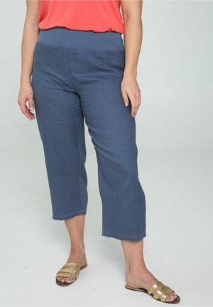 Vrouw draagt losse blauwe korte broek, koraalkleurige top en gouden platte sandalen, staand op een lichte vloer tegen een effen achtergrond.
