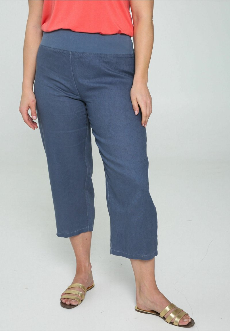 Femme portant un pantalon bleu ample et court, un haut corail et des sandales plates dorées, debout sur un sol clair devant un fond uni.