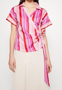 Chemise à manches courtes, style portefeuille, présentant des rayures verticales dans des nuances de rose et de rouge. Le tissu semble léger ; comprend un détail noué à la taille.