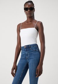 White fitted tank top with thin straps paired with high-waisted blue denim jeans. Model wearing black cat-eye sunglasses and hoop earrings.