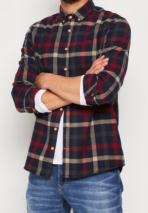 Hombre con los brazos cruzados que lleva una camisa de cuadros roja, azul marino y beige abotonada sobre una camiseta blanca y pantalones vaqueros azules.