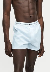 Buste masculin portant un boxer Tommy Hilfiger bleu clair avec une ceinture marquée, debout devant un fond clair uni.