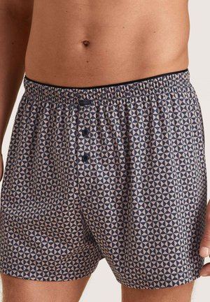 Marineblauwe boxers met een geometrisch crèmekleurig patroon, elastische tailleband en twee knopen aan de voorkant. Gladde stoftextuur.