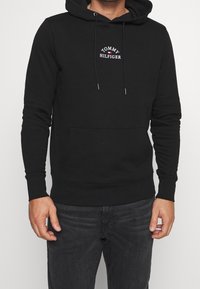 Black hoodie made of cotton, features a front pocket, drawstring hood, and a logo embroidered in white and red. Smooth texture, fitted design.